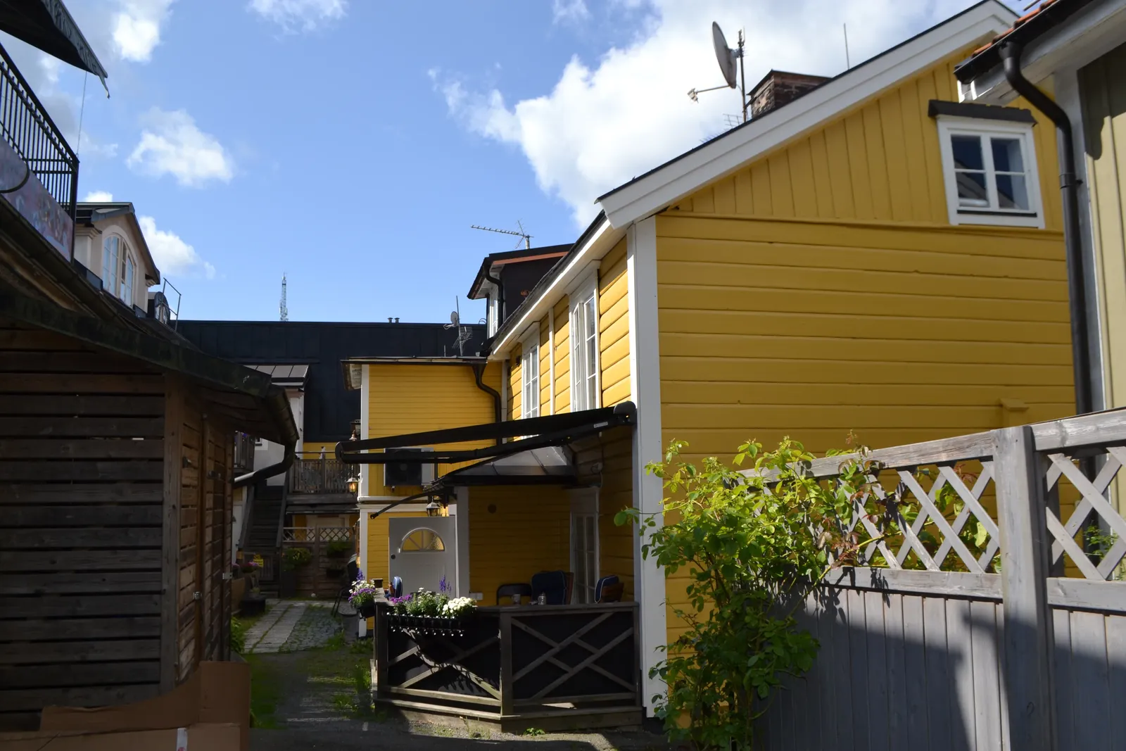 Smal gård mellan gula trähus, med en liten trätrall med blommor och ett vitt staket med rutmönster.
