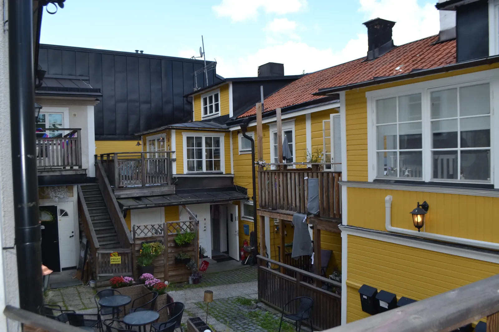 Gårdsrum sett uppifrån med gula trähus, trappor, balkonger, blomlådor och uteserveringsmöbler på gatstenen.