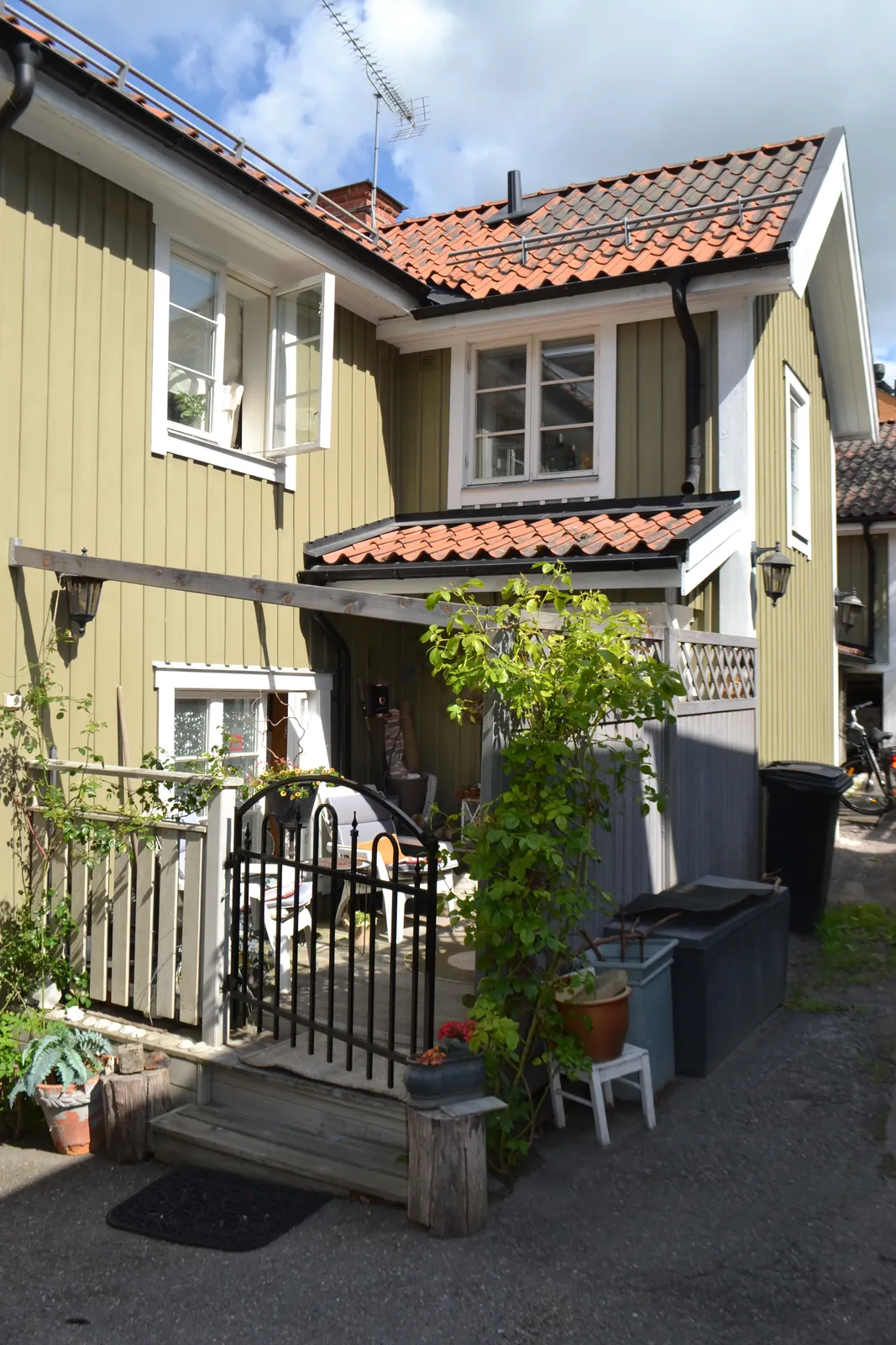 Trähus med tegeltak, liten veranda med järngrind och en uppvuxen klätterväxt, parkerad container och grusyta framför.