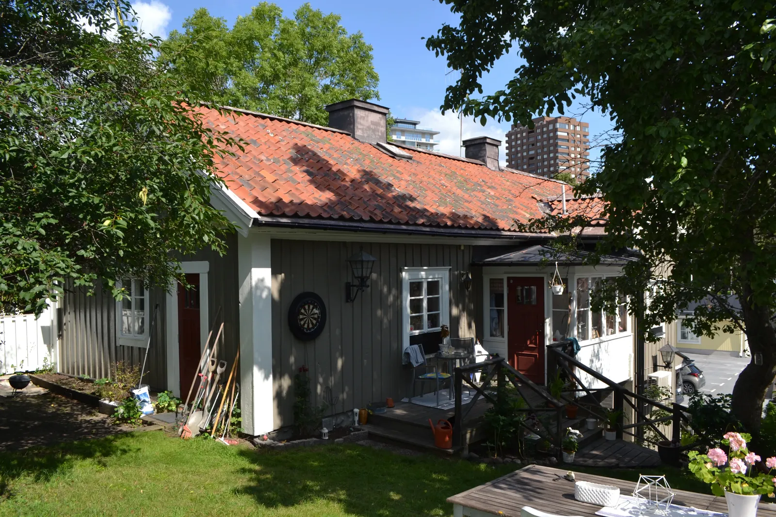Låg grå trähusbyggnad med tegeltak, röda dörrar, en veranda och en trädgård med gräsmatta.